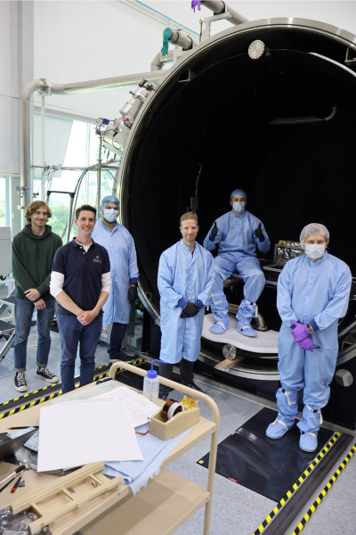 A group of people stand in front of a Thermal and Vacuum Space Simulation Chamber at ANU's Advanced Instrumentation and Technology Centre. The two men to the left are dressed in business casual, the two men in the centre are dressed in blue lab coats and the two men to the right are dressed in full blue clean-room attire. 